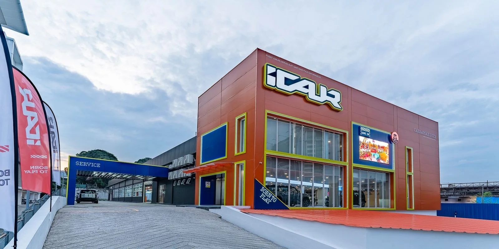An iCAUR 4S centre at dusk — orange and yellow signage above the showroom glass,
              with a covered service lane running down the left side